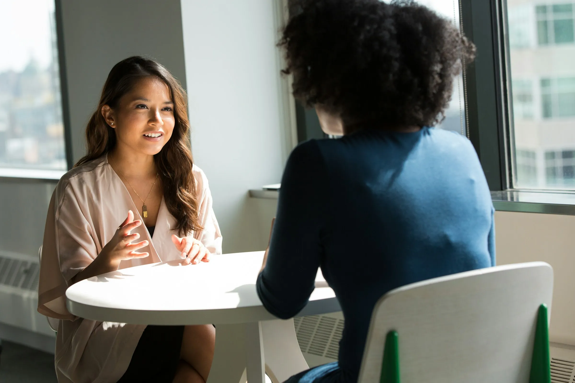 Two women talking across a round table by a window